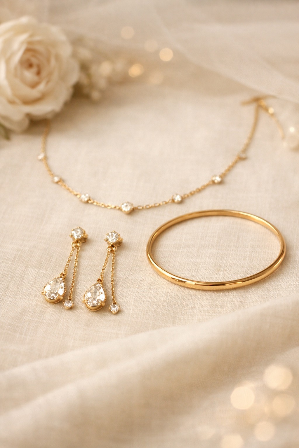 A gold station necklace, pear-drop earrings and a plain gold bangle laid on ivory linen beside a white rose